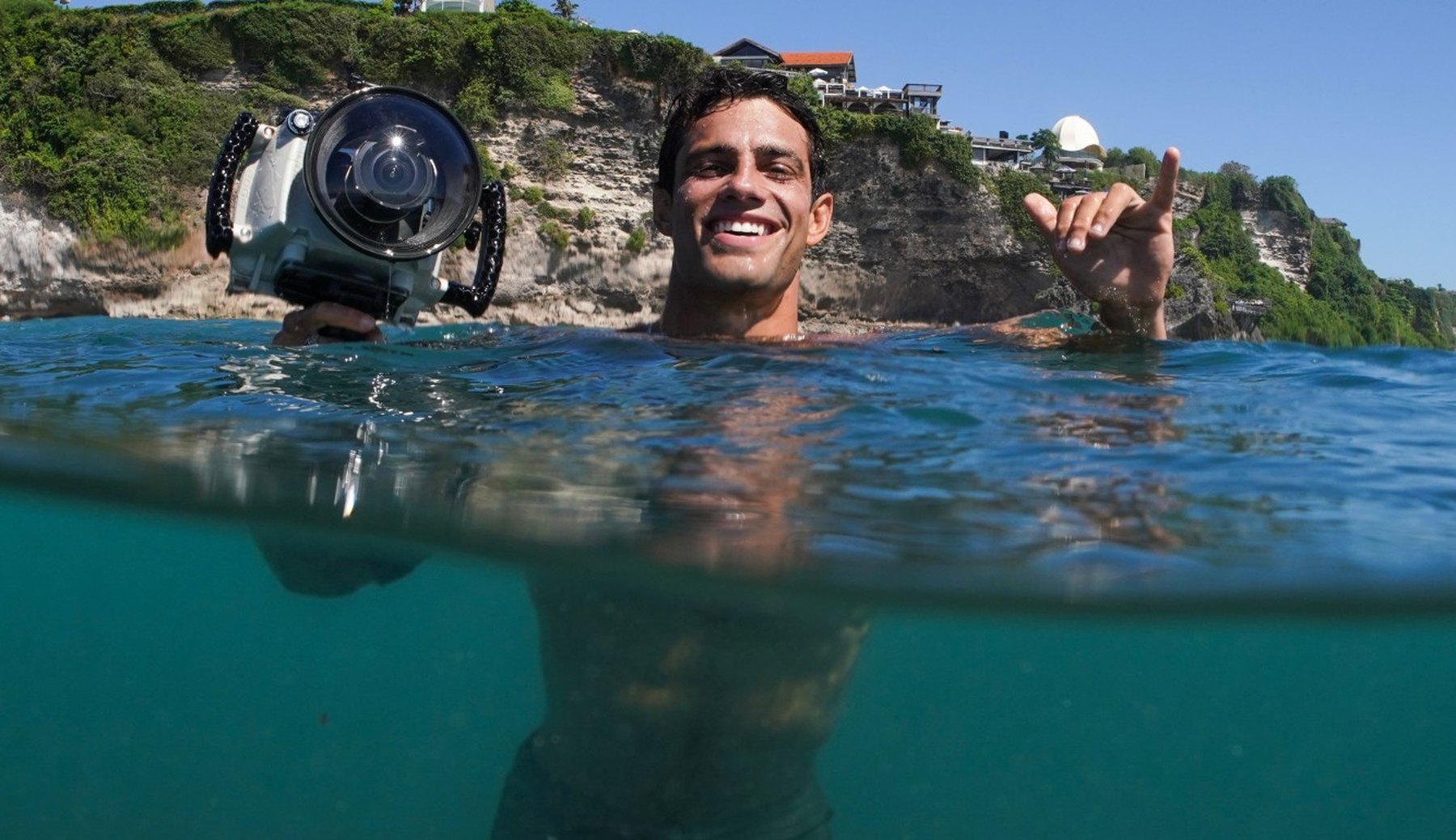 Rafael Fragoso smiling in the water at Uluwatu with an underwater camera housing, the cliff behind him
